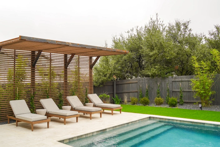 Four tan lounge chairs sit under a custom pergola by a pool.
